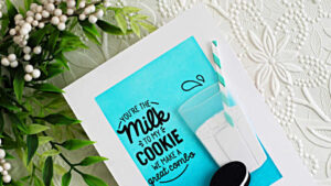 A handmade card featuring a white A2 card base with a bright turquoise ink-blended panel in the center. A bold black sentiment reads, “You’re the Milk to My Cookie — We Make a Great Combo.” To the right is a dimensional die-cut glass of milk with a striped straw, and along the bottom are three Oreo-style sandwich cookies. The card is photographed on a white embossed floral background with greenery in the upper corner and color swatches partially visible to the side.