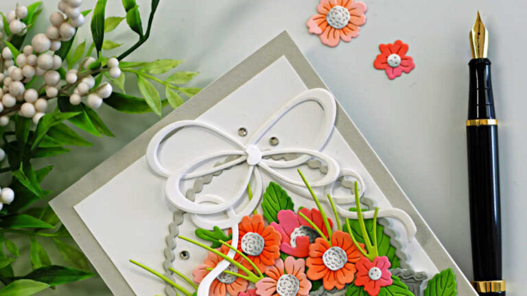 Handmade Easter card featuring the Country Basket Bouquet Die and Double Loop Bow from the Simon Says Stamp Beautiful Moments release. A textured die-cut basket filled with coral, peach, and pink flowers with layered green leaves sits on a white panel layered over a Fog cardstock card base. A large white double loop bow tops the basket, while small gems add sparkle and a “Happy Easter” sentiment completes the cheerful spring design.