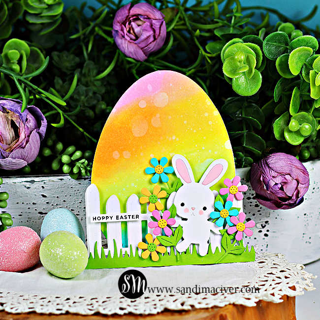 Easter egg-shaped card with rainbow ink blending, die cut bunny, flowers, and fence, styled with pastel eggs and spring greenery