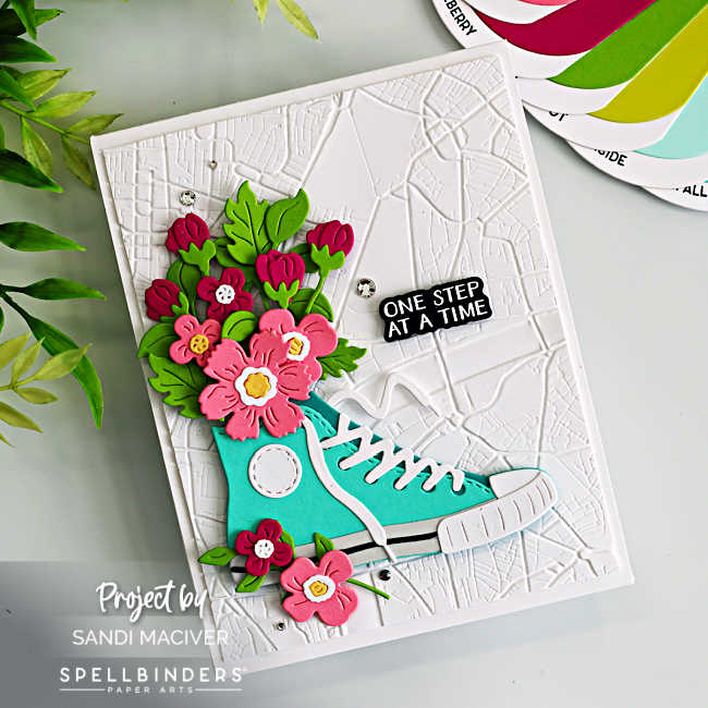 Handmade A2 card featuring the Spellbinders Classic Kicks Large Die of the Month (March 2026). A teal die-cut sneaker with white laces sits on a white background embossed with the Urban Grid 3D Embossing Folder of the Month. The sneaker is filled with layered pink and red flowers and bright green leaves spilling from the top. A small black sentiment strip reading “One Step at a Time,” from the Good Times Clear Stamp and Die of the Month, is placed to the right, with a few silver gems scattered for sparkle.