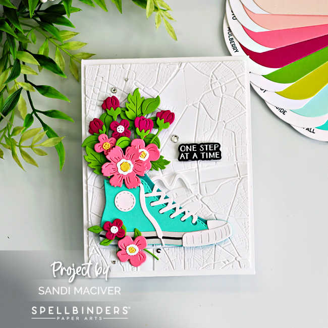 Handmade A2 card featuring the Spellbinders Classic Kicks Large Die of the Month (March 2026). A teal die-cut sneaker with white laces sits on a white background embossed with the Urban Grid 3D Embossing Folder of the Month. The sneaker is filled with layered pink and red flowers and bright green leaves spilling from the top. A small black sentiment strip reading “One Step at a Time,” from the Good Times Clear Stamp and Die of the Month, is placed to the right, with a few silver gems scattered for sparkle.