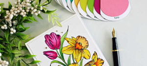 Handmade spring floral card featuring bright pink tulips and yellow daffodils created with the Spellbinders Yana’s Tulips BetterPress and Stencil Set. The flowers are colored with coordinating layering stencils and Prismacolor pencils on a white card panel and finished with a black and gold “Sending a bouquet of hugs” sentiment.