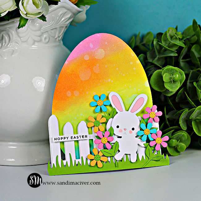 Easter egg-shaped card with rainbow ink blending, die cut bunny, colorful flowers, and white fence, displayed standing upright