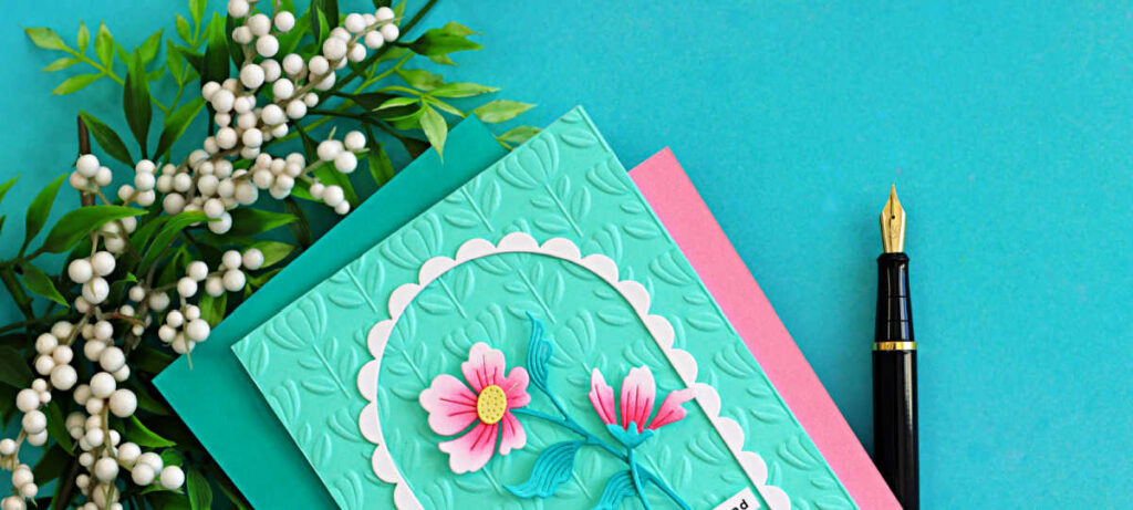 Elegant floral arch handmade card featuring pink die cut flowers on an aqua embossed background with scalloped frame, styled with greenery and a fountain pen