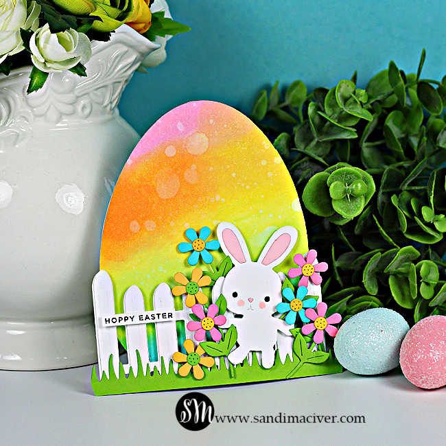 Rainbow Easter egg-shaped card with die cut bunny, flowers, and white fence, displayed with pastel eggs and greenery