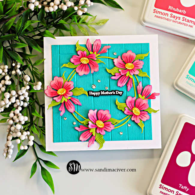 Square Mother’s Day card with bright pink die cut flowers arranged in a wreath on Audrey Blue cardstock embossed with a wood plank texture, featuring a “Happy Mother’s Day” sentiment and scattered gems.