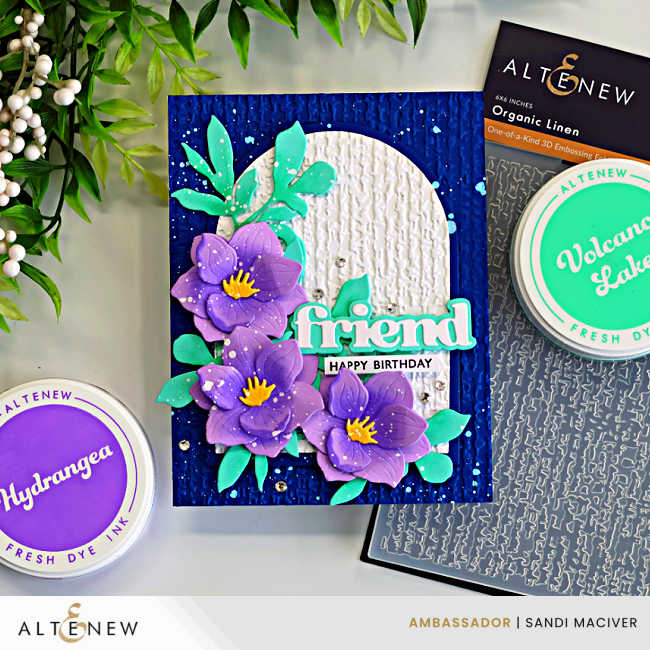 Magnolia die cut card with purple layered flowers, teal leaves, and embossed linen arch on a navy background, featuring Altenew Hydrangea and Volcano Lake inks and Organic Linen embossing folder