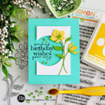 Handmade floral birthday card featuring a circle die cut window with stamped “sending birthday wishes your way” sentiment, layered yellow flowers, teal card base, and green gems, surrounded by cardmaking supplies