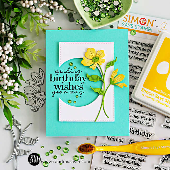 Handmade floral birthday card featuring a circle die cut window with stamped “sending birthday wishes your way” sentiment, layered yellow flowers, teal card base, and green gems, surrounded by cardmaking supplies