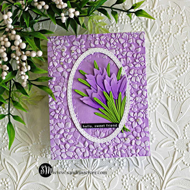 Crocus die cut card with layered purple flowers and green leaves inside a stitched oval frame on a lilac embossed floral background, finished with a “hello, sweet friend” sentiment and small gems.