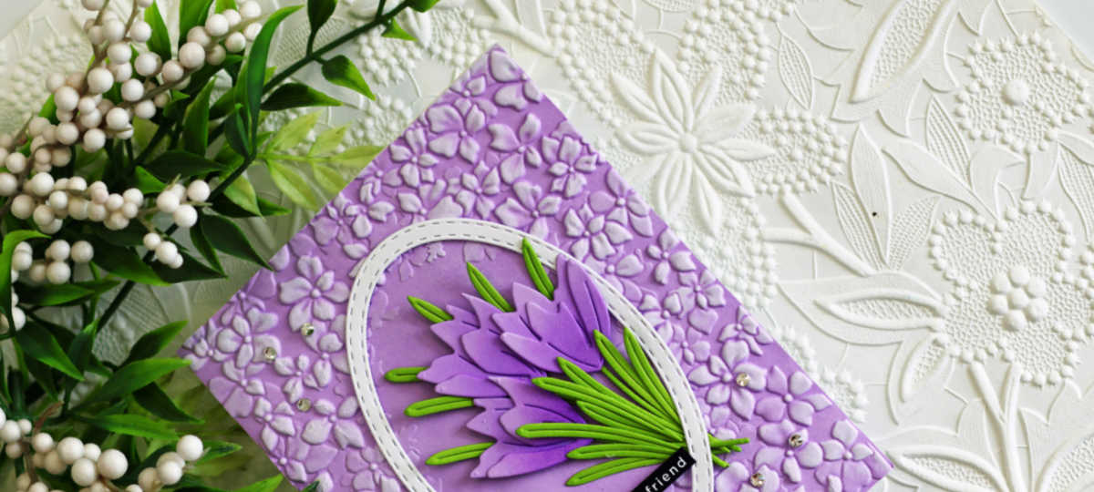 Purple crocus die cut card featuring layered flowers and green leaves inside a stitched oval frame, set on an embossed floral background with added gems for dimension.
