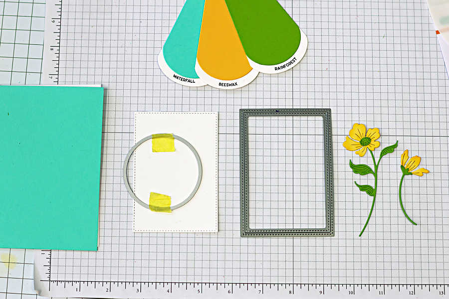 Cardmaking supplies layout showing a circle die positioned on a white panel for a circle die cut card, with teal, yellow, and green cardstock, stitched rectangle die, and yellow floral die cuts ready for assembly