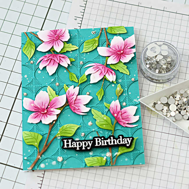 Finished die cut spring card with pink blossom branches on a teal embossed background and Happy Birthday sentiment with gems.