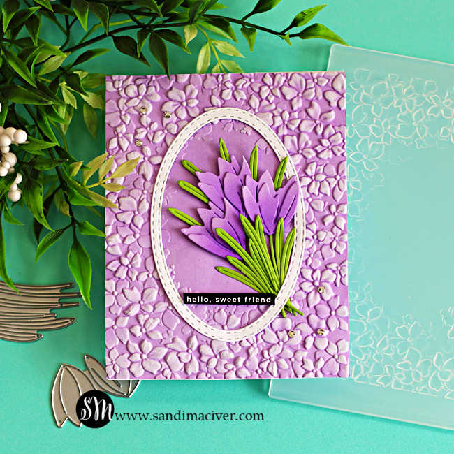 Crocus die cut card with layered purple flowers and green leaves inside a stitched oval frame on a lilac embossed floral background, with stencil and dies shown on a teal workspace.