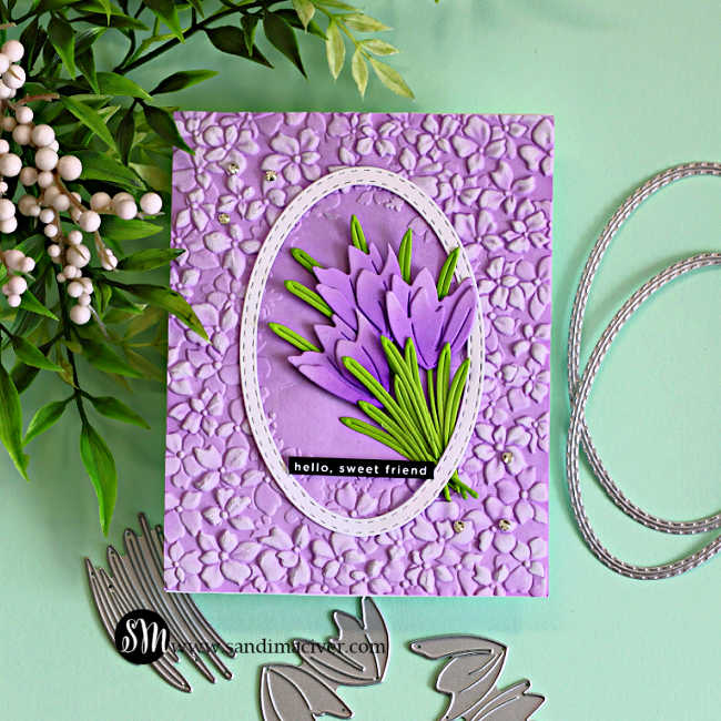 Crocus die cut card featuring layered purple flowers and green leaves inside a stitched oval frame on a lilac embossed floral background, with coordinating metal dies displayed on a teal surface.