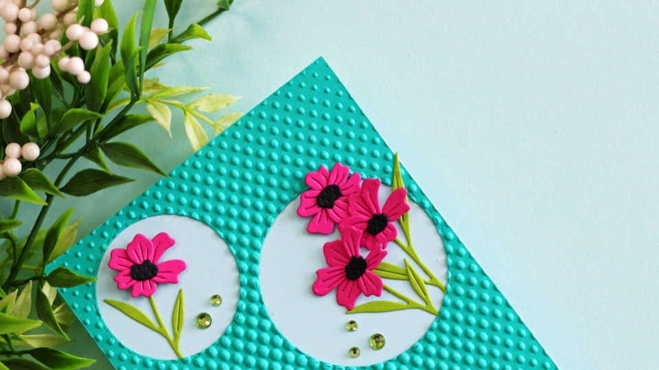 Close-up of a handmade card featuring spotlighting die cut florals inside stitched circle frames on a teal embossed background with halftone dots, showcasing bright pink flowers and green leaves.