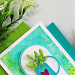 Watering can birthday card with ink smooshed Distress Oxide background and stenciled texture, featuring green die cut foliage on a clean white card base.