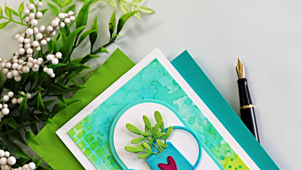 Watering can birthday card with ink smooshed Distress Oxide background and stenciled texture, featuring green die cut foliage on a clean white card base.