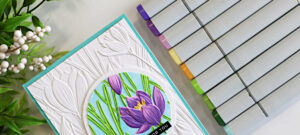 Handmade spotlight technique card featuring the Simon Says Stamp Crocus Field embossing folder with a circular crocus floral focal point colored using purple and green Copic markers on a white embossed background with aqua card base.