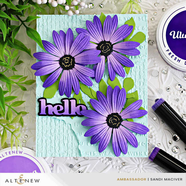 Handmade Altenew card featuring three layered purple African daisies with embossed petals and black centers arranged over a soft aqua mixed-media background with embossed texture, distressed details, bright green leaves, and a bold “hello” sentiment.