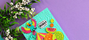 Colorful tropical die cut card featuring a bright parrot, citrus slices, watermelon, and umbrella created with the Spellbinders May Large Die of the Month. The aqua embossed background from the Radiant Day 3D Embossing Folder adds sunny texture to this Caribbean color splash summer card design.