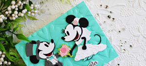 Close-up of a handmade wedding card featuring die cut classic mouse bride and groom characters from Spellbinders on a mint green embossed heart background. The bride holds a pink floral bouquet while sparkling gems and a white embossed floral background add elegant texture and shine.