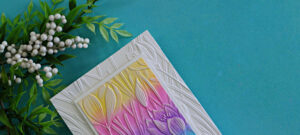 Handmade card featuring an embossing folder background with a die cut frame, ink blended panel in soft rainbow colors, and white ink highlighting the raised floral details
