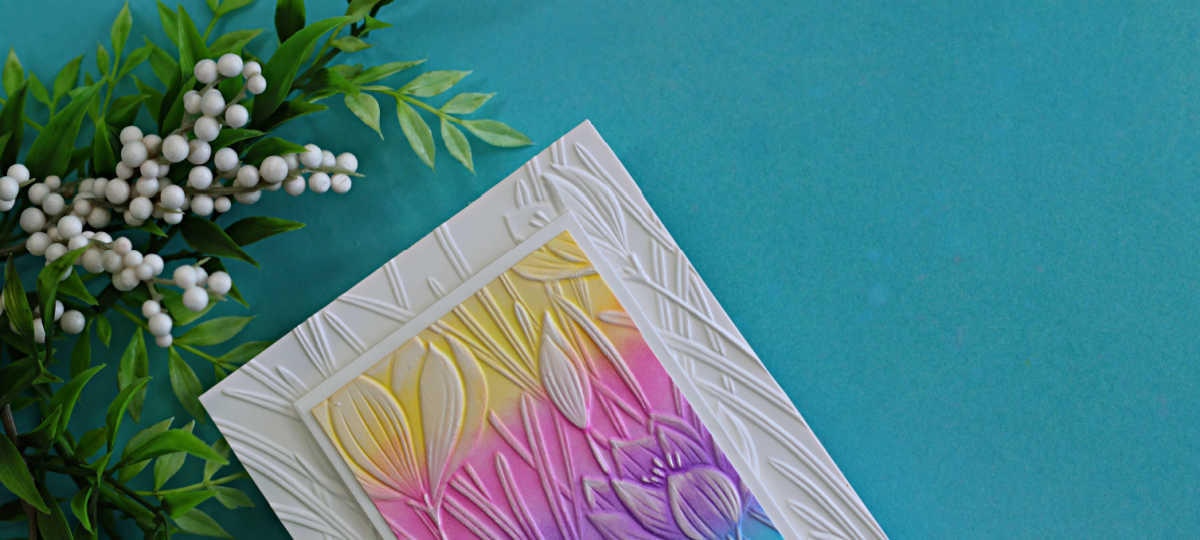 Handmade card featuring an embossing folder background with a die cut frame, ink blended panel in soft rainbow colors, and white ink highlighting the raised floral details