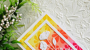 Close-up of a handmade Hello Spring card featuring bright yellow, orange, and pink retro daisy stencil blending with layered white Arctic Poppies die cuts and a dimensional white frame on a textured floral background.