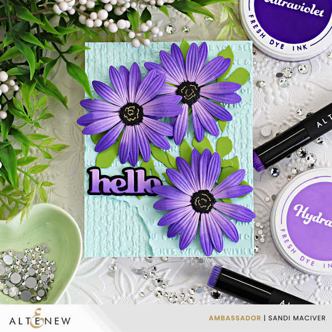 Handmade Altenew card featuring three layered purple African daisies with embossed petals and black centers arranged over a soft aqua mixed-media background with embossed texture, distressed details, bright green leaves, and a bold “hello” sentiment.
