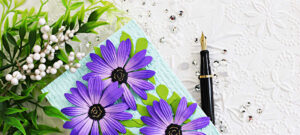 Close-up of a handmade card featuring layered purple African daisies with embossed petals and black centers on a soft aqua textured background. The mixed-media card design includes distressed embossed layers, bright green leaves, sparkling gems, and a black fountain pen styled on a white embossed floral backdrop.