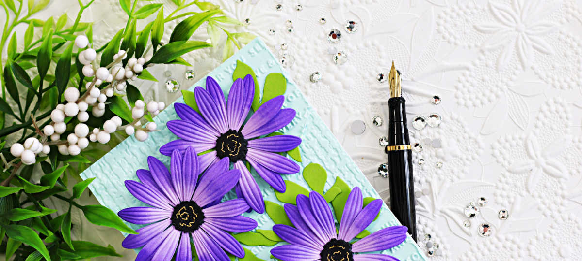 Close-up of a handmade card featuring layered purple African daisies with embossed petals and black centers on a soft aqua textured background. The mixed-media card design includes distressed embossed layers, bright green leaves, sparkling gems, and a black fountain pen styled on a white embossed floral backdrop.
