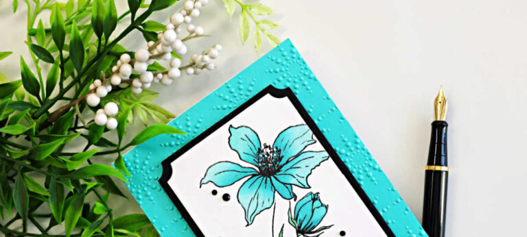 Close-up of a handmade floral card featuring turquoise blue flowers from the Wild and Free stamp set framed with black and white A2 Notched Rectangle die cuts on a bright turquoise embossed background, photographed with greenery and a black fountain pen.