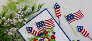 Close-up of a patriotic die cut card featuring layered American flags, fireworks rockets, and red, white, and blue floral blooms arranged in a star-spangled hat. The background is embossed with the Spellbinders Night Sky Celebration embossing folder for added texture and detail.