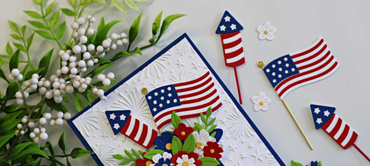 Close-up of a patriotic die cut card featuring layered American flags, fireworks rockets, and red, white, and blue floral blooms arranged in a star-spangled hat. The background is embossed with the Spellbinders Night Sky Celebration embossing folder for added texture and detail.