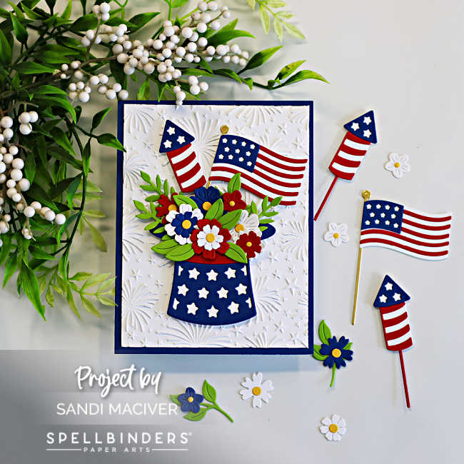 Patriotic handmade card featuring layered die cut American flags, fireworks rockets, and red, white, and blue flowers arranged inside a star-spangled hat. The card background is embossed with the Spellbinders Night Sky Celebration embossing folder for added texture and festive detail.