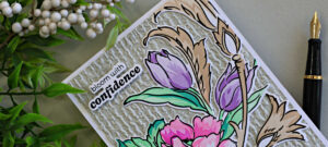 Close-up of a handmade floral card featuring layered pink flowers, lavender tulips, aqua green leaves, and warm beige flourishes over a textured ivory embossed background. The card includes the sentiment “bloom with confidence” and is styled with greenery and a black fountain pen.