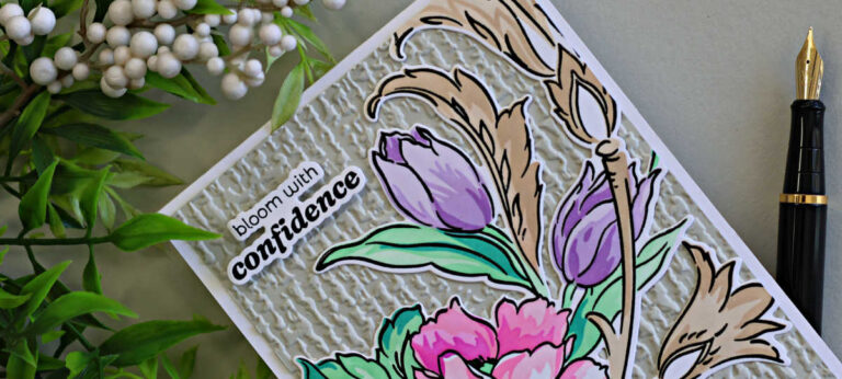 Close-up of a handmade floral card featuring layered pink flowers, lavender tulips, aqua green leaves, and warm beige flourishes over a textured ivory embossed background. The card includes the sentiment “bloom with confidence” and is styled with greenery and a black fountain pen.