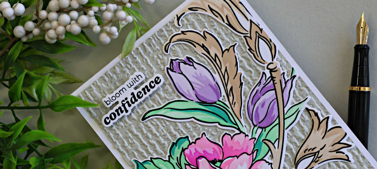 Close-up of a handmade floral card featuring layered pink flowers, lavender tulips, aqua green leaves, and warm beige flourishes over a textured ivory embossed background. The card includes the sentiment “bloom with confidence” and is styled with greenery and a black fountain pen.