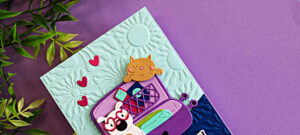 Close-up of a whimsical travel-themed handmade card featuring a purple suitcase filled with a puppy and kitten, created with the Spellbinders Ready to Roll Small Die of the Month. The card includes a soft aqua embossed background using the Radiant Day 3D Embossing Folder, layered die cuts, and bright pink heart accents.