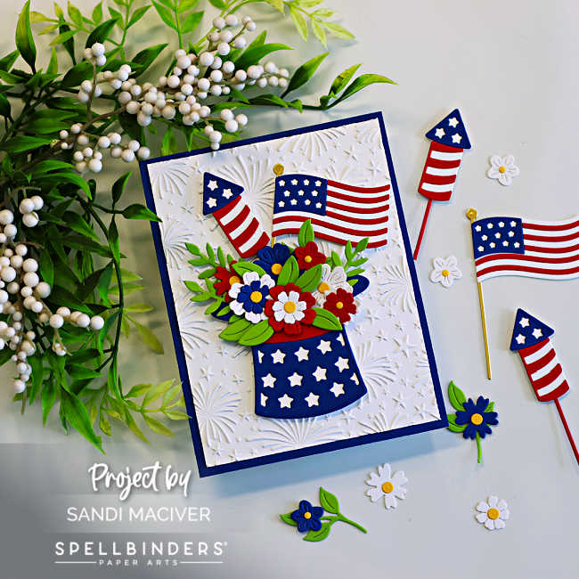 Patriotic handmade card featuring layered die cut American flags, fireworks rockets, and red, white, and blue flowers arranged inside a star-spangled hat. The card background is embossed with the Spellbinders Night Sky Celebration embossing folder for added texture and festive detail.