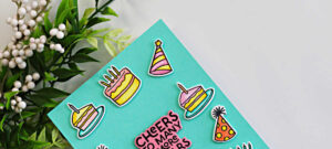 Close-up of a simple die cut birthday card featuring Hero Arts My Monthly Hero Yah Day images, with colorful cakes and party hats on a teal background and a bold “Cheers to Many More Years” sentiment.