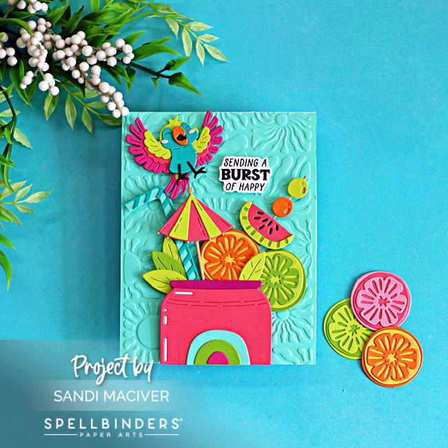 Bright tropical summer card featuring colorful citrus die cuts, a tropical parrot, watermelon slice, and umbrella arranged in a pink drink cup on an aqua embossed background. Created with the Spellbinders May Large Die of the Month and Radiant Day 3D Embossing Folder in a bold Caribbean color palette.