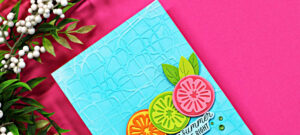 Close-up of a bright summer handmade card featuring layered citrus die cuts in pink, lime, and orange on a turquoise embossed background created with the Spellbinders May Embossing Folder Club.