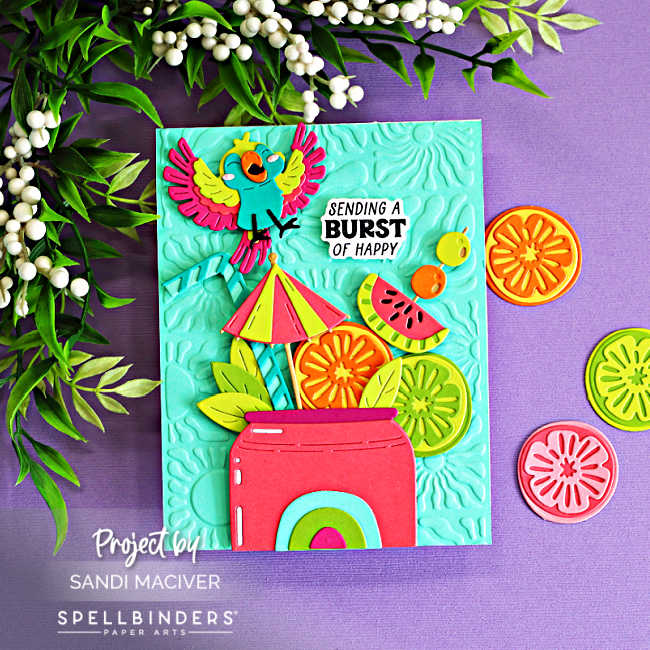 Tropical summer die cut card featuring a colorful parrot, citrus slices, watermelon, and umbrella arranged in a bright pink drink cup. Created with the Spellbinders May Large Die of the Month and Radiant Day 3D Embossing Folder using bold Caribbean-inspired colors on an aqua embossed background.