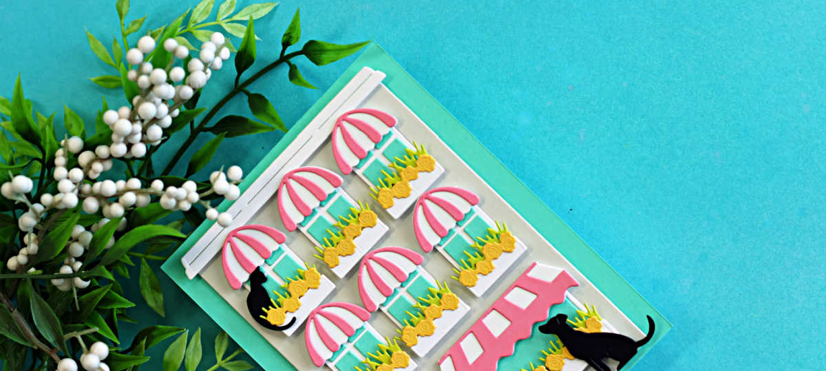 Close up of a layered die cut storefront card created with the Spellbinders Yana’s Everyday City Holiday dies featuring pink striped awnings, flower-filled window boxes, black cats, and a soft spring color palette of Teal Topaz, Waterfall, Peridot, Tutu, Fog, and black cardstock.