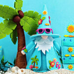 Handmade tropical Fergus felt gnome decor featuring a turquoise gnome wearing pink sunglasses beside a coordinating surfboard. The scene includes a palm tree, seashells, sandy beach accents, and lush greenery, creating a fun summer vacation display in bright aqua, pink, and yellow tones.