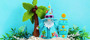 Handmade tropical Fergus felt gnome decor featuring a turquoise gnome wearing pink sunglasses beside a coordinating surfboard. The scene includes a palm tree, seashells, sandy beach accents, and lush greenery, creating a fun summer vacation display in bright aqua, pink, and yellow tones.