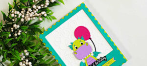 Close-up of a handmade birthday card featuring a cute lime green and lavender dragon holding a bright pink balloon. The dragon is framed inside a teal polaroid-style die-cut window with a white embossed background and yellow scalloped accents. Green foliage with white berries decorates the left side of the image, creating a cheerful and whimsical birthday card design.