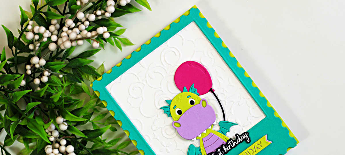 Close-up of a handmade birthday card featuring a cute lime green and lavender dragon holding a bright pink balloon. The dragon is framed inside a teal polaroid-style die-cut window with a white embossed background and yellow scalloped accents. Green foliage with white berries decorates the left side of the image, creating a cheerful and whimsical birthday card design.